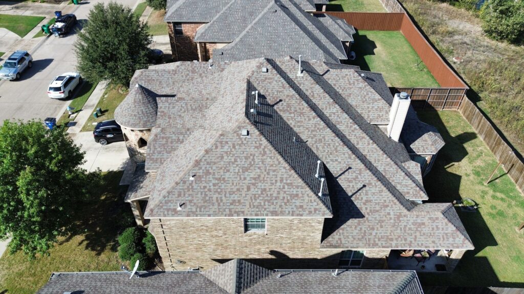 Overhead view of a complex gray architectural-shingle hip roof with a turret and neighboring roofs in a Celina, Texas neighborhood