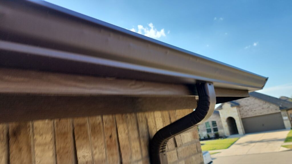 Close-up of a brown seamless gutter and downspout on a tan brick home in Celina, Texas