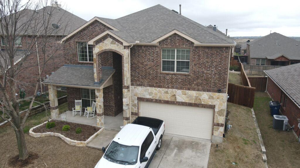 Aerial view of a two-story brick home with a stone-column porch, white garage door, and gray architectural-shingle gables in Celina, Texas