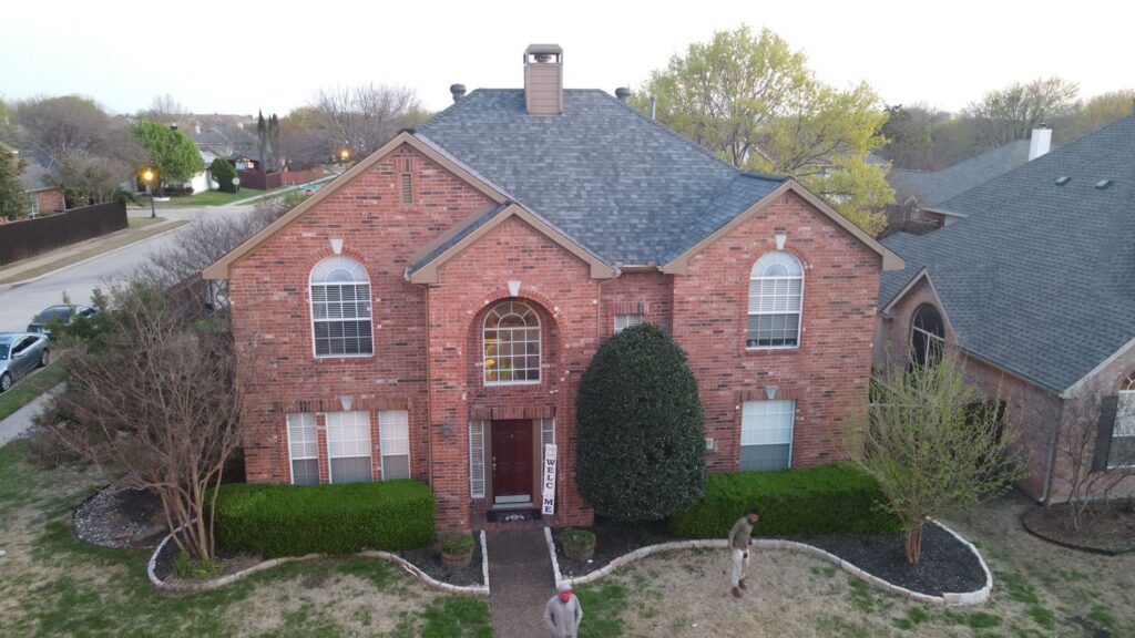 Aerial view of a two-story red brick Allen, Texas home with a completed gray architectural-shingle hip roof and circular front walk