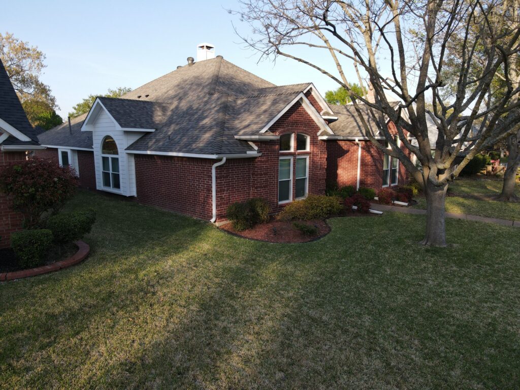 Aerial view of a one-story red brick ranch with a completed gray architectural-shingle hip roof, white chimney, and side-yard lawn in Allen, Texas
